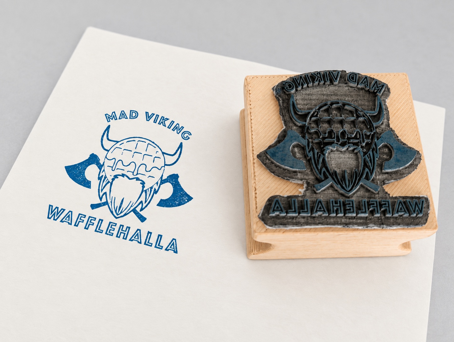 Enhanced product photograph of the Mad Viking Wafflehalla wood-mounted stamp beside its blue printed impression